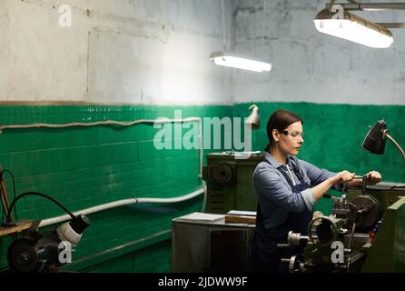 Concentrated middle-aged brunette woman in safety goggles performing turning operation on lathe in dirty factory shop Stock Photo