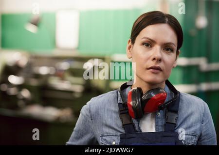 Portrait of serious attractive middle-aged female factory worker with ear protectors on neck standing in industrial shop Stock Photo