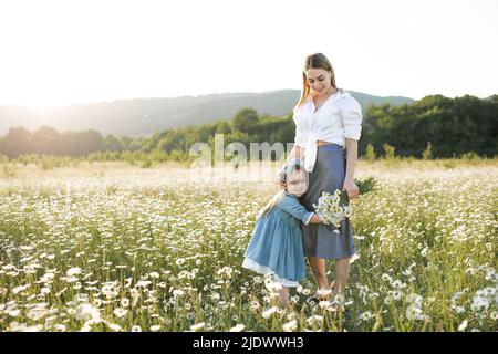 Pretty woman holding kid girl 3-4 year old standing in camomile meadow over nature background outdoor. Mother with little child together. Motherhood. Stock Photo