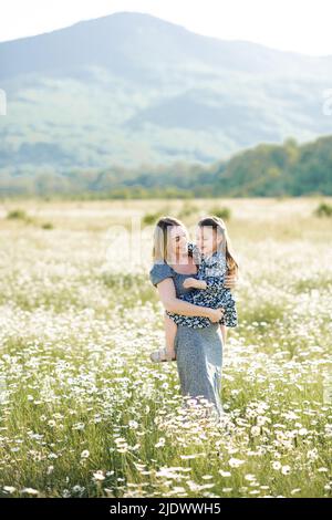 Cute smiling woman holding baby girl daughter 3-4 year old wear similar dresses posing in blooming floral meadow outdoor. Mother toddler in field over Stock Photo