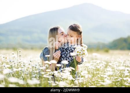 Cute mother kissing child girl daughter in meadow with blooming flowers outdoor together. Family lifestyle. Motherhood. Summer season. Stock Photo