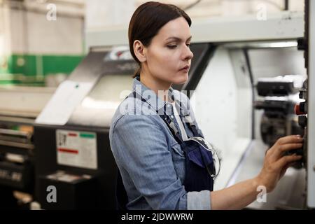 Serious busy middle-aged brunette woman standing in factory workshop and running cnc lathe Stock Photo