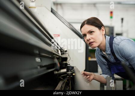 Content skilled brunette woman in workwear watching lathe machine processing and adjusting it at factory Stock Photo