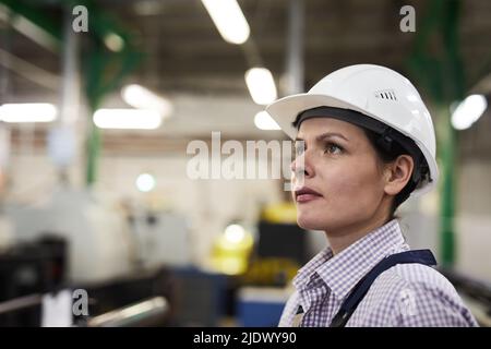 Serious purposeful young female engineer in white hardhat standing in workshop of industrial plant and looking forward Stock Photo