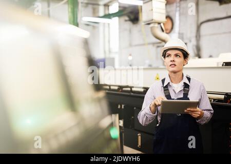 Introspective experienced young woman in hardhat standing at factory and using digital tablet while analyzing cnc lathe processes Stock Photo