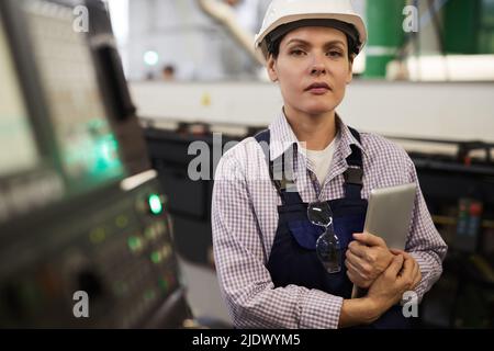 Portrait of serious confident female industrial engineer in hardhat and workwear standing at cnc lathe and holding tablet Stock Photo