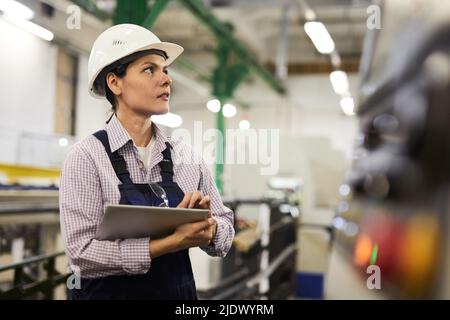Serious thoughtful female technical engineer in hardhat standing at factory and using tablet for operating cnc machine Stock Photo
