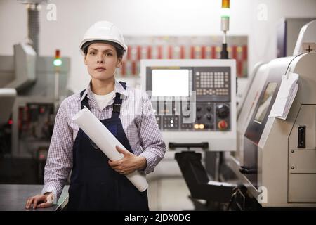 Portrait of serious confident female engineer in hardhat standing at cnc machine and holding rolled blueprint Stock Photo