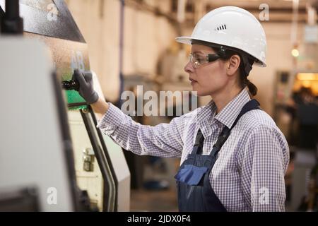 Concentrated gritty young woman in hardhat and safety goggles preparing detail for processing and closing cover of cnc machine at factory Stock Photo