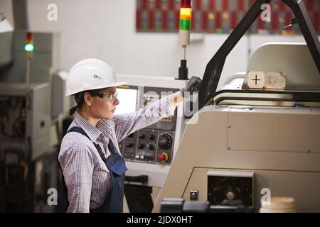 Serious experienced female CNC machine operator standing at lathe and preparing for work at factory Stock Photo
