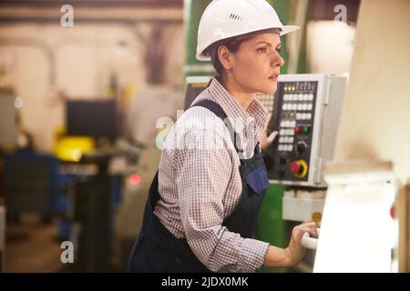 Serious modern female engineer in work helmet standing at factory and controlling work of lathe Stock Photo