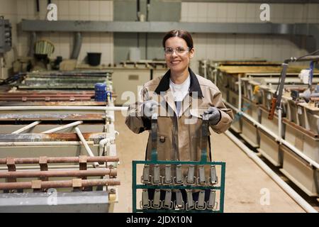 Portrait of cheerful young brunette woman in protective eyewear standing at factory and carrying metal belt parts in frame Stock Photo
