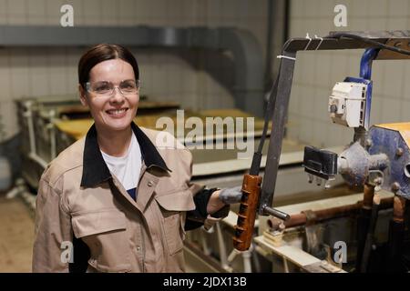 Happy attractive middle-aged woman in workwear standing at assembly line in industrial area and pushing button on control panel Stock Photo
