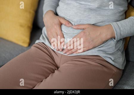 Sick young woman suffering from menstrual pain. Woman with hands squeezing belly having painful stomach ache or period cramps sitting on sofa Stock Photo