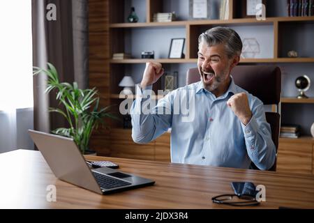 Happy businessman surprised by the good news and making yes gesture while looking at laptop sitting at office. Trader enjoying success, celebrating Stock Photo