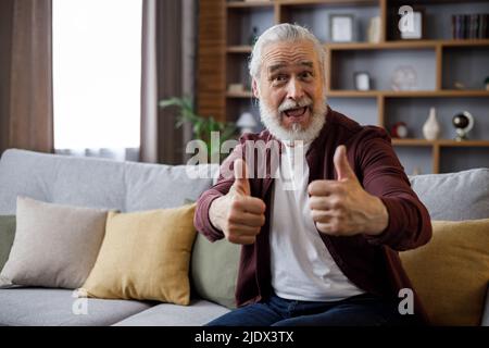 Smiling older man sitting on couch giving thumb up while playing computer game, looking at camera, smiling. Stock Photo