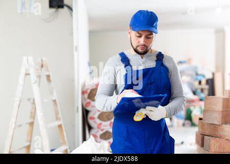 Responsible focused bearded supervisor wearing blue overall taking notes on papers while inspecting construction site Stock Photo