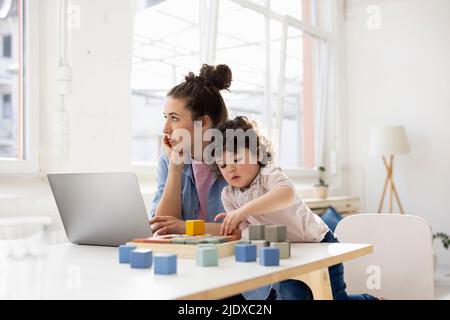 Mother working from home using laptop while daughter is playing with buiilding blocks Stock Photo