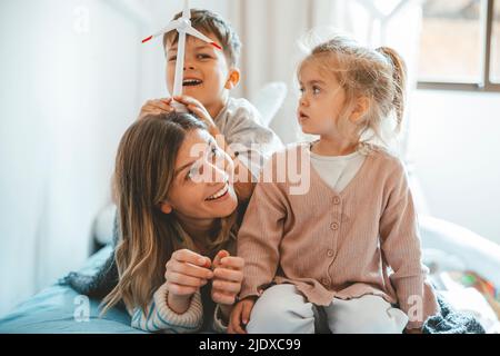 Playful boy holding wind turbine on mother's head sitting by sister at home Stock Photo