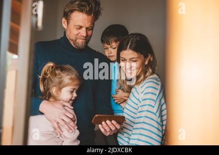 Smiling man sharing smart phone with family seen through window Stock Photo