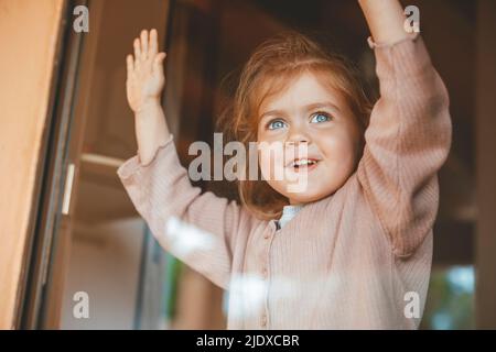 Happy cute girl at home looking out through glass window Stock Photo