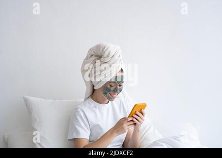 Happy woman wearing facial mask using smart phone in bedroom Stock Photo
