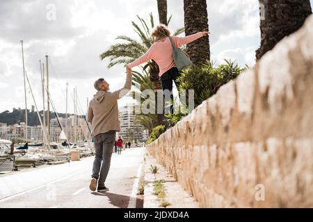 Mature man holding hand of woman walking on wall in city Stock Photo