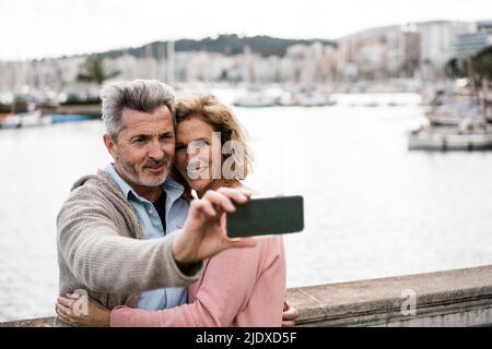 Smiling woman embracing man taking selfie in front of sea Stock Photo