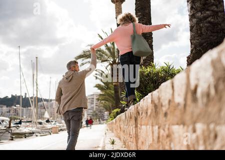 Mature man holding hand of woman walking on wall by harbor Stock Photo