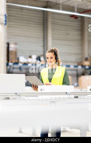 Worker wearing headset holding tablet PC examining box on conveyor belt in warehouse Stock Photo