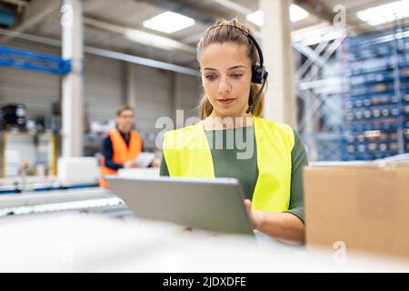 Young worker wearing headset using tablet PC by cardboard box in warehouse Stock Photo
