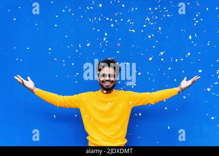 Smiling man with arms outstretched standing under falling confetti in front of blue wall Stock Photo