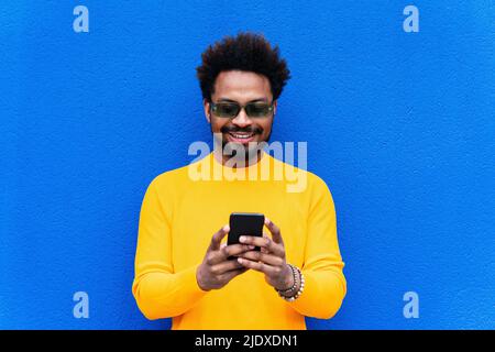 Smiling Afro man using smart phone in front of blue wall Stock Photo