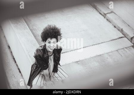 Cheerful young woman with Afro hairstyle sitting on wall Stock Photo