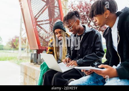 Smiling young man using laptop sitting by friends on wall Stock Photo