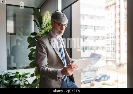 Businessman with documents using tablet PC standing by window in office Stock Photo