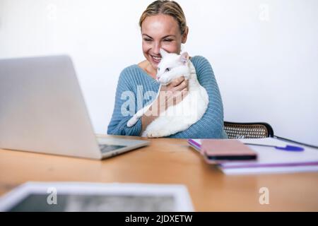 Smiling businesswoman with pet cat looking at laptop working from home Stock Photo
