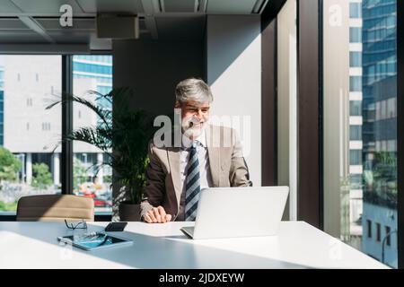 Happy businessman using laptop sitting by window at desk in office Stock Photo