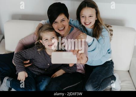 Smiling mother taking selfie with daughters sitting on sofa at home Stock Photo