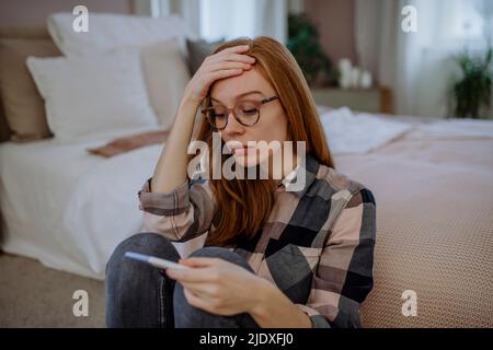 Worried woman with pregnancy testing kit sitting in front of bed at home Stock Photo