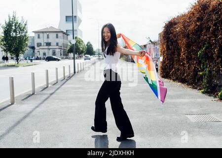 Cheerful young woman with rainbow flag walking on road Stock Photo
