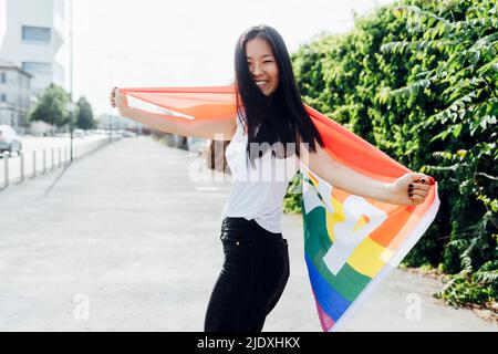 Happy woman with arms outstretched standing with rainbow flag on sunny day Stock Photo