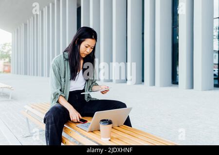 Young freelancer using tablet PC sitting with laptop and reusable coffee on bench Stock Photo