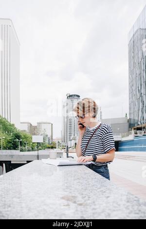 Architect talking on smart phone looking at blueprint standing by wall Stock Photo