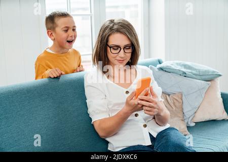 Happy boy looking at mother using smart phone on sofa at home Stock Photo
