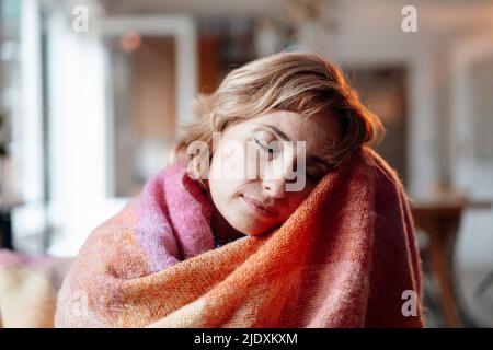 Woman wrapped in blanket sitting with eyes closed at home Stock Photo
