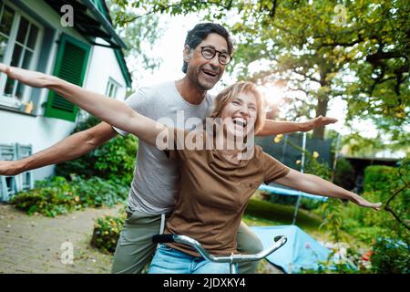 Cheerful mature woman and man with arms outstretched on bicycle in back yard Stock Photo