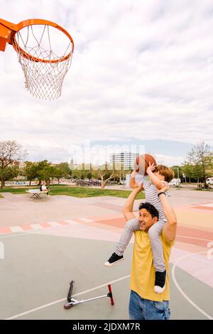Boy with basketball sitting on father's shoulders at sports court Stock Photo