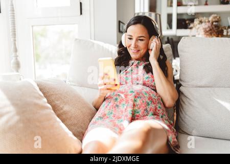 Smiling pregnant woman listening music through wireless headphones using smart pone sitting on sofa at home Stock Photo