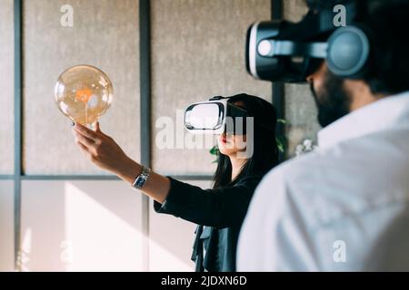Businesswoman with VR glasses examining light bulb by colleague at office Stock Photo